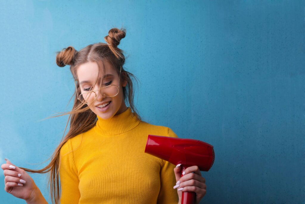 Women with round glasses, hair buns and bright yellow shirt is holding a hair dryer to iron a shirt 