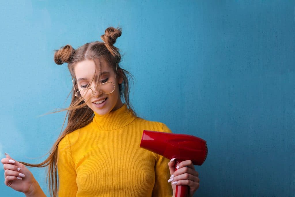 Women with round glasses, hair buns and bright yellow shirt is holding a hair dryer to iron a shirt