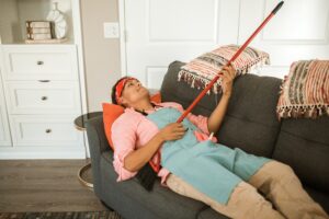 Man Wearing a Pink Long Sleeves and Blue Apron Holding a Broom While Lying Down on a Cou