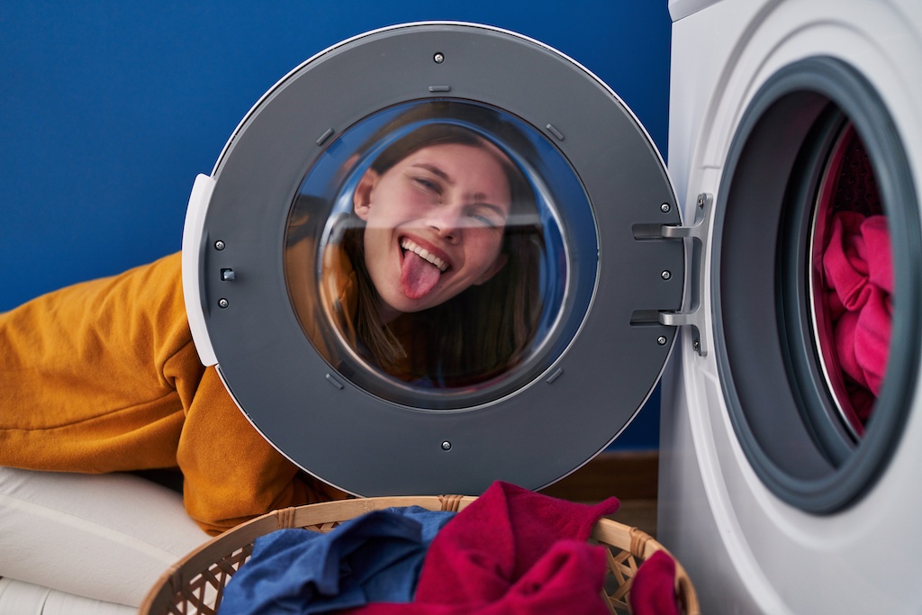 Young brunette woman looking through the washing machine window sticking tongue out happy with funny expression.